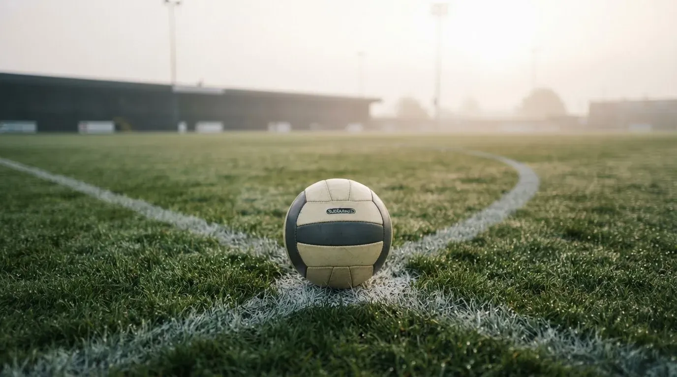 Ein einzelner Fussball liegt auf dem Anstosspunkt eines leeren Rasenplatzes am frühen Morgen, Tau glitzert auf dem Gras, die Tribüne liegt im Dunst.