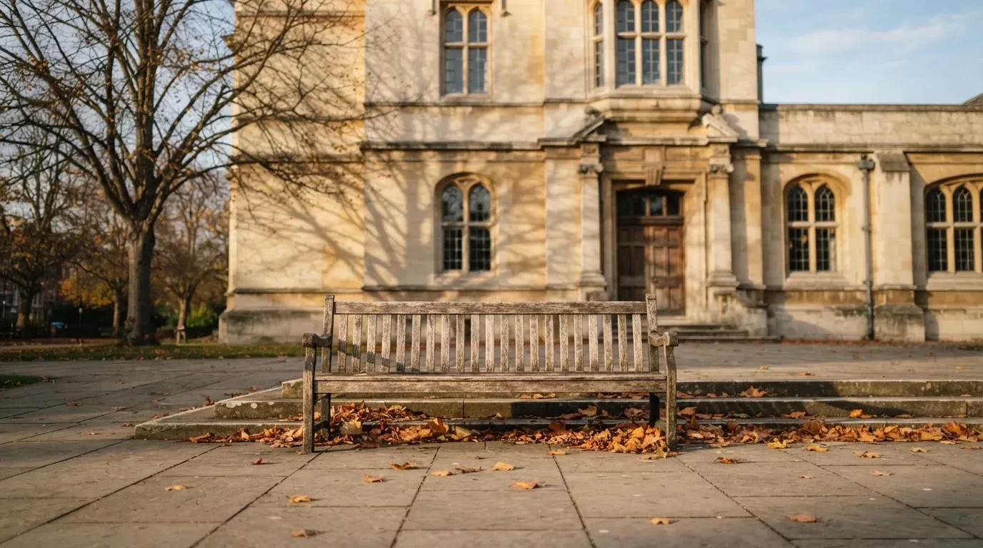 Eine Holzbank steht vor einem Verwaltungsgebäude aus Stein mit hohen Fenstern, Herbstlaub liegt auf dem Boden.