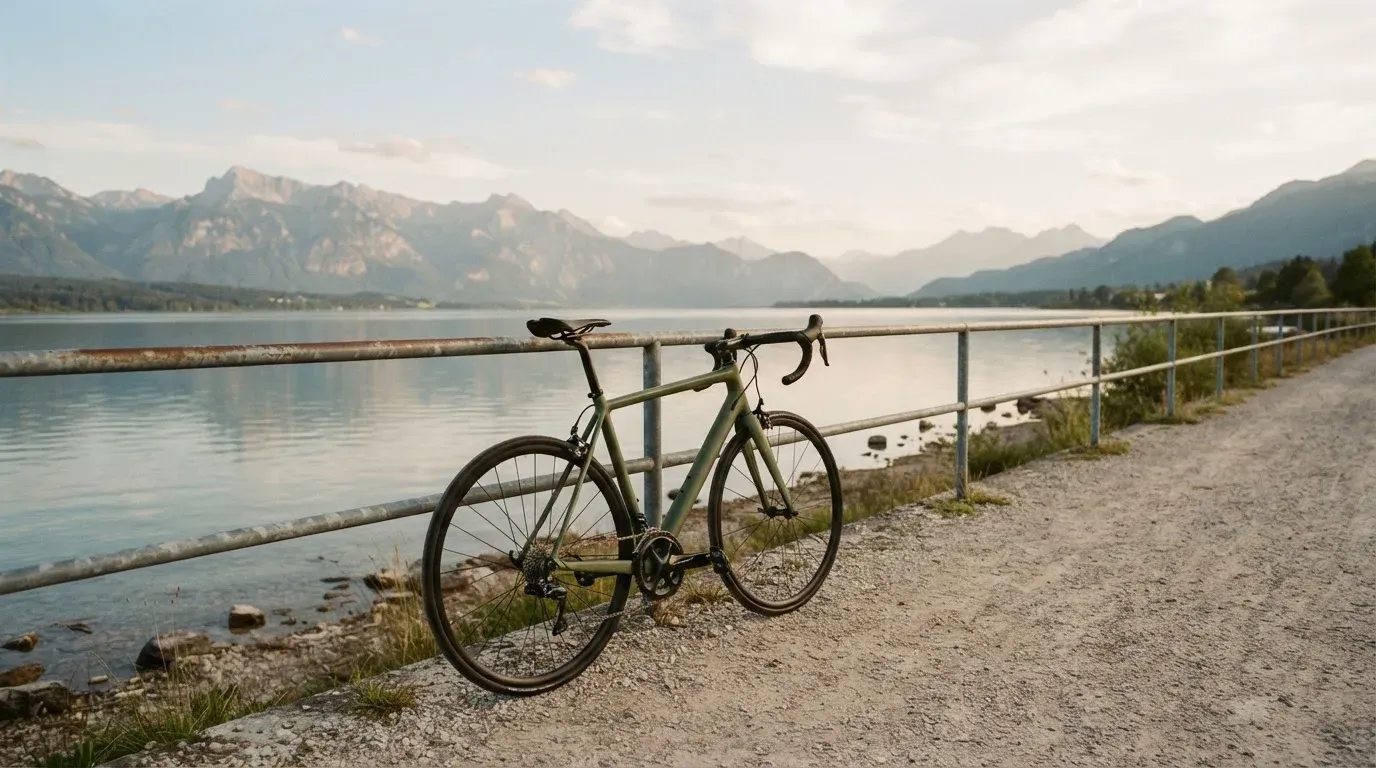 Ein Rennrad lehnt an einem Geländer am Seeufer, dahinter liegt ruhiges Wasser vor einer Bergkette.