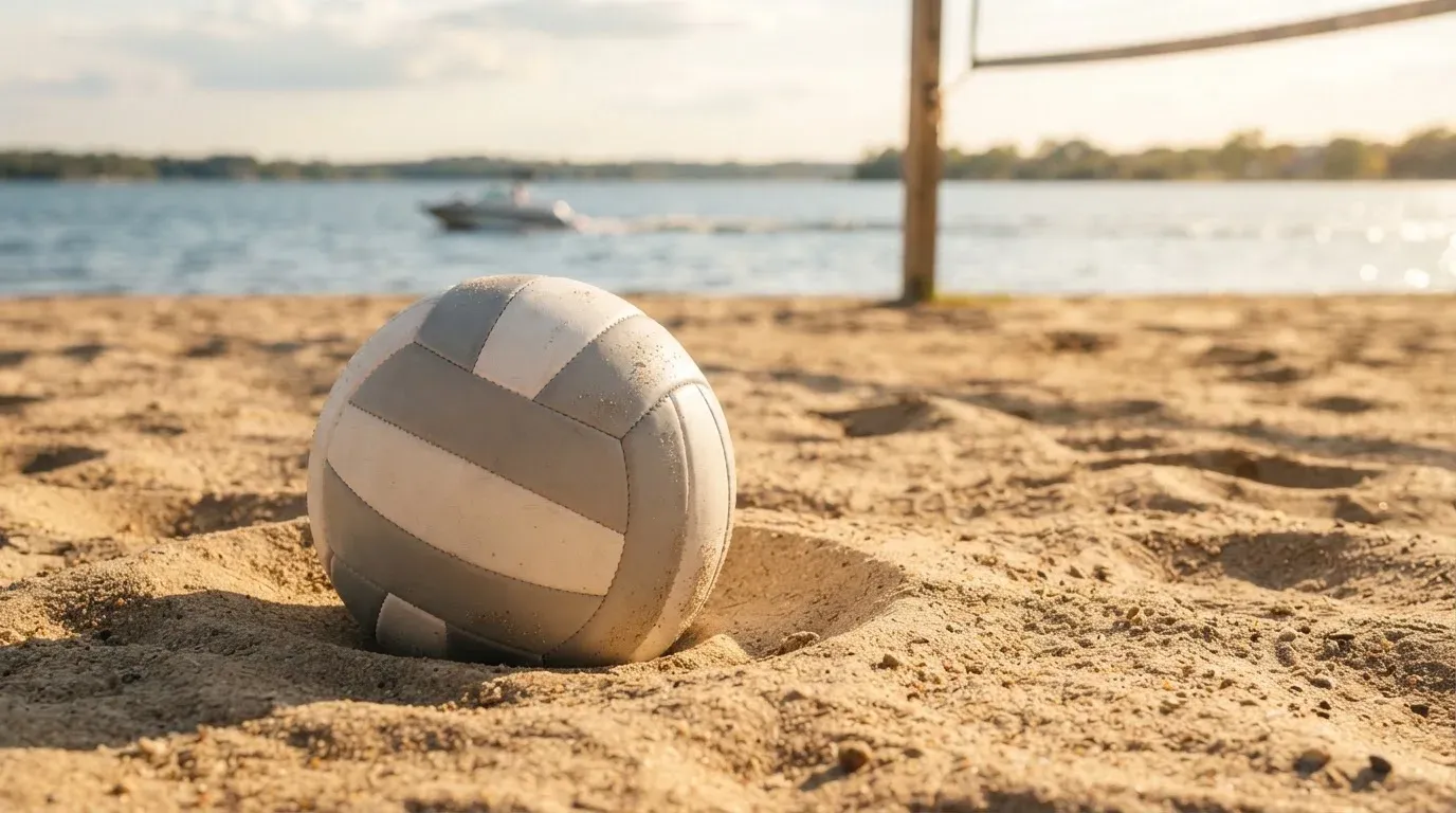 Ein Volleyball liegt im Sand eines Spielfelds am Seeufer, im Hintergrund gleitet ein Boot über das Wasser.