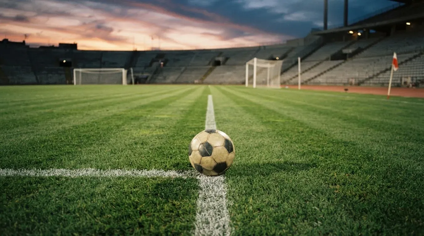 Ein Fussball liegt auf dem Mittelkreis eines leeren Rasenplatzes, dahinter ragen leere Tribünenreihen in den Abendhimmel.