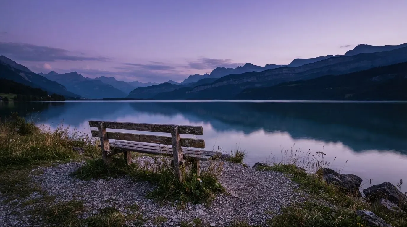 Eine Holzbank steht in der Dämmerung am Seeufer, im Hintergrund zeichnet sich ein Bergpanorama gegen den violetten Himmel ab.