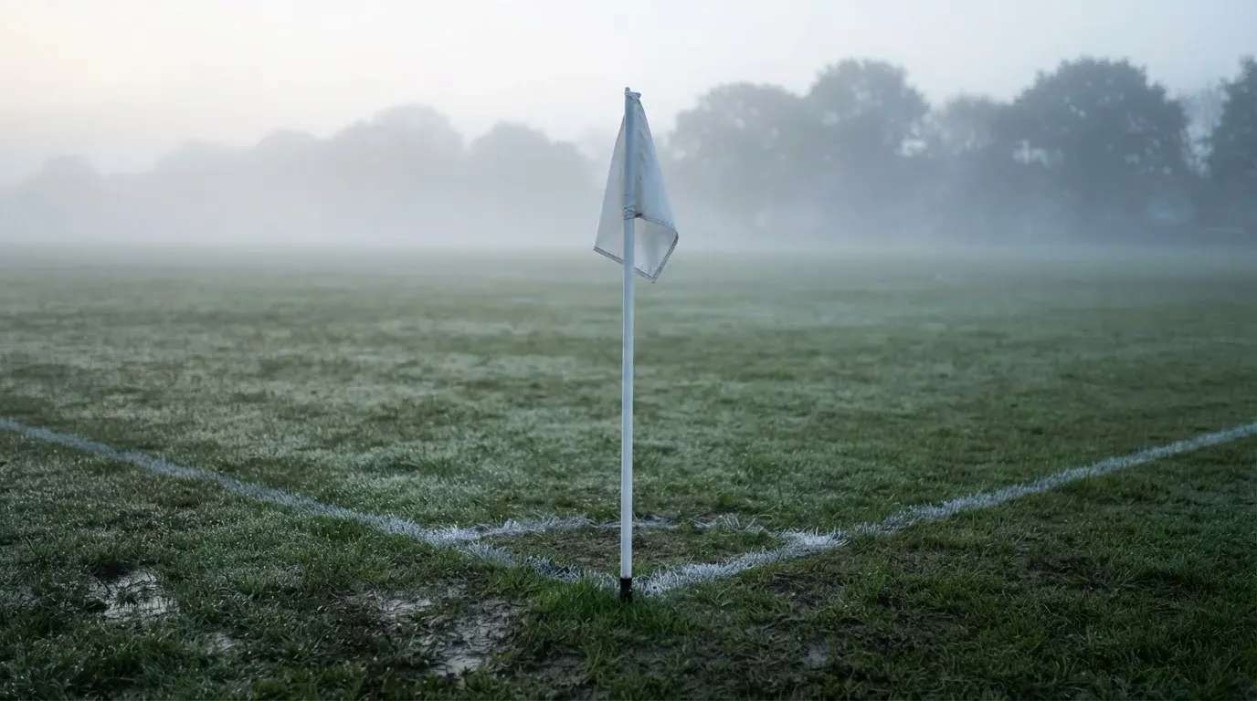Eine Eckfahne steht am Morgen auf nassem Rasen, Tautropfen hängen im Gras und über dem Platz liegt leichter Dunst.