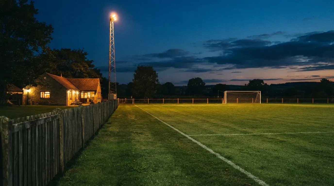 Ein kleiner Dorf-Fussballplatz liegt in der Dämmerung unter einem hohen Flutlichtmast, am Spielfeldrand steht ein Holzzaun.