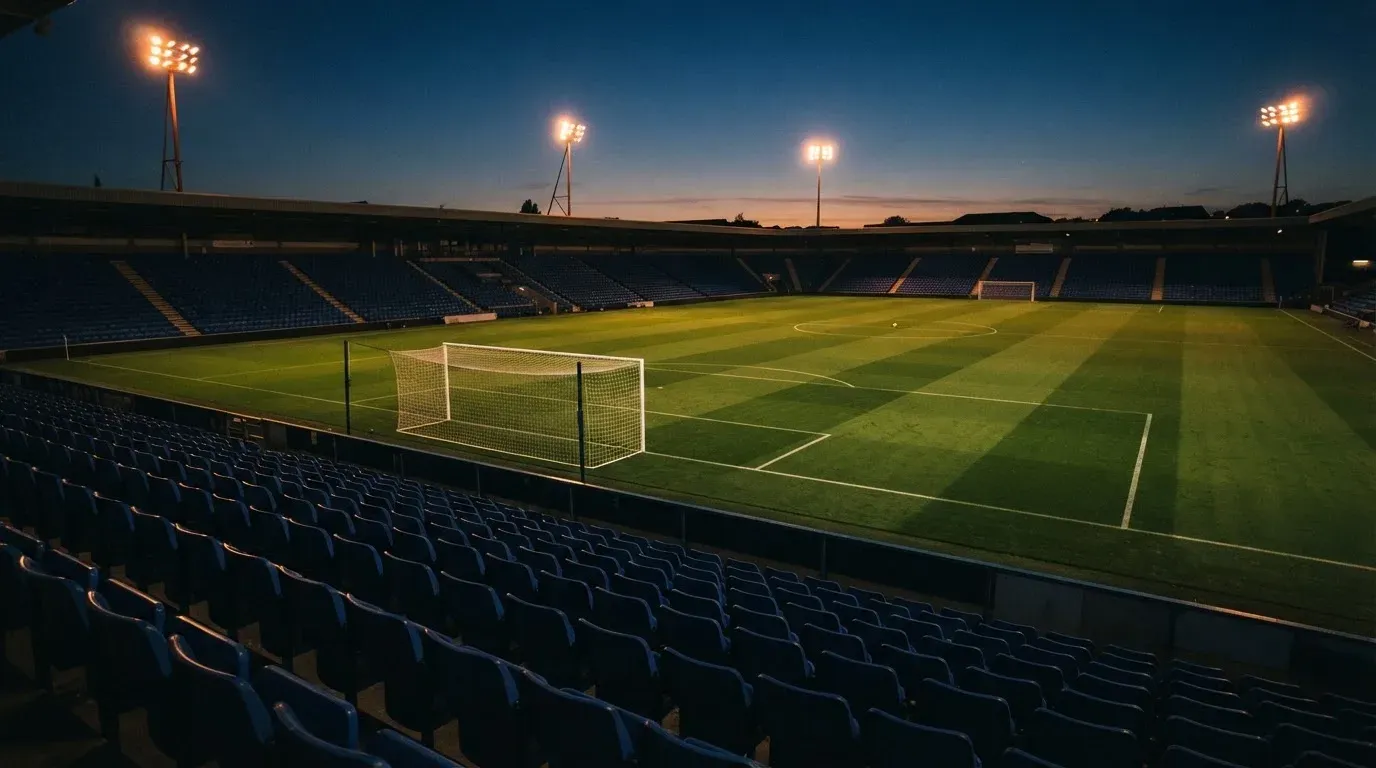 Leere Tribünenränge eines Fussballstadions in der Abenddämmerung, über dem Rasen leuchten die Flutlichter.