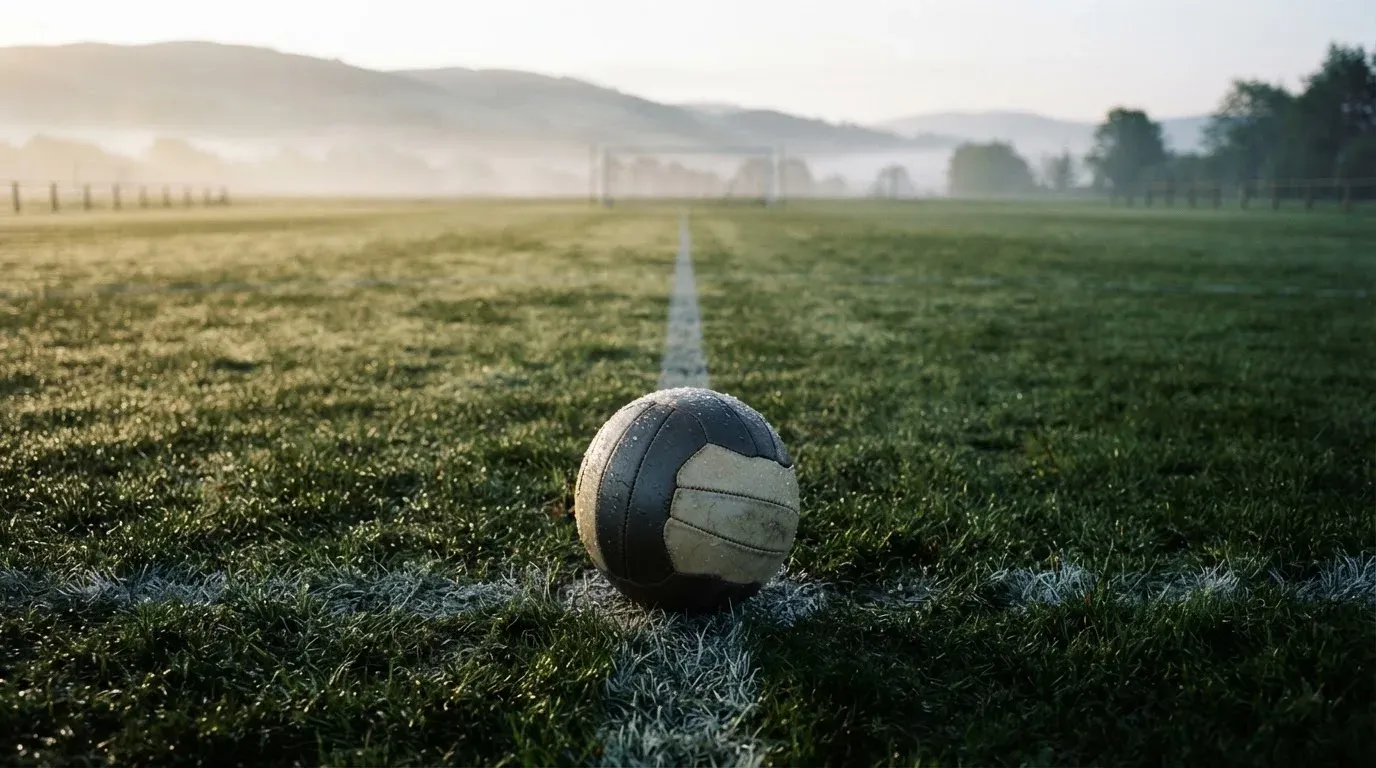 Ein Fussball liegt am frühen Morgen auf dem Anstosspunkt eines taunassen Rasens, im Hintergrund erheben sich Hügel im Dunst.