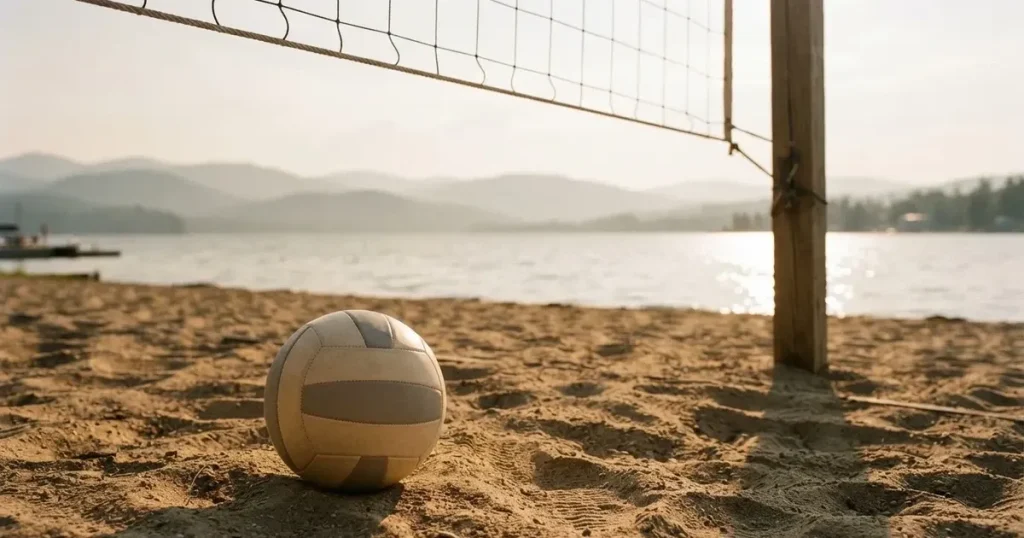 Ein Volleyball liegt am Fuss des Netzes auf Sand, im Hintergrund glitzert ein See und ein Bergzug steht im Dunst.