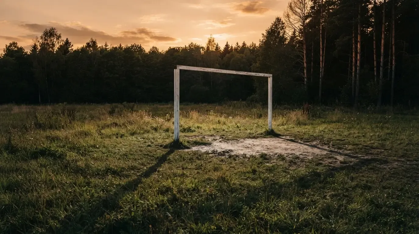 Ein Fussballtor ohne Netz steht auf einer Wiese am Waldrand, die Abenddämmerung färbt den Himmel orange.