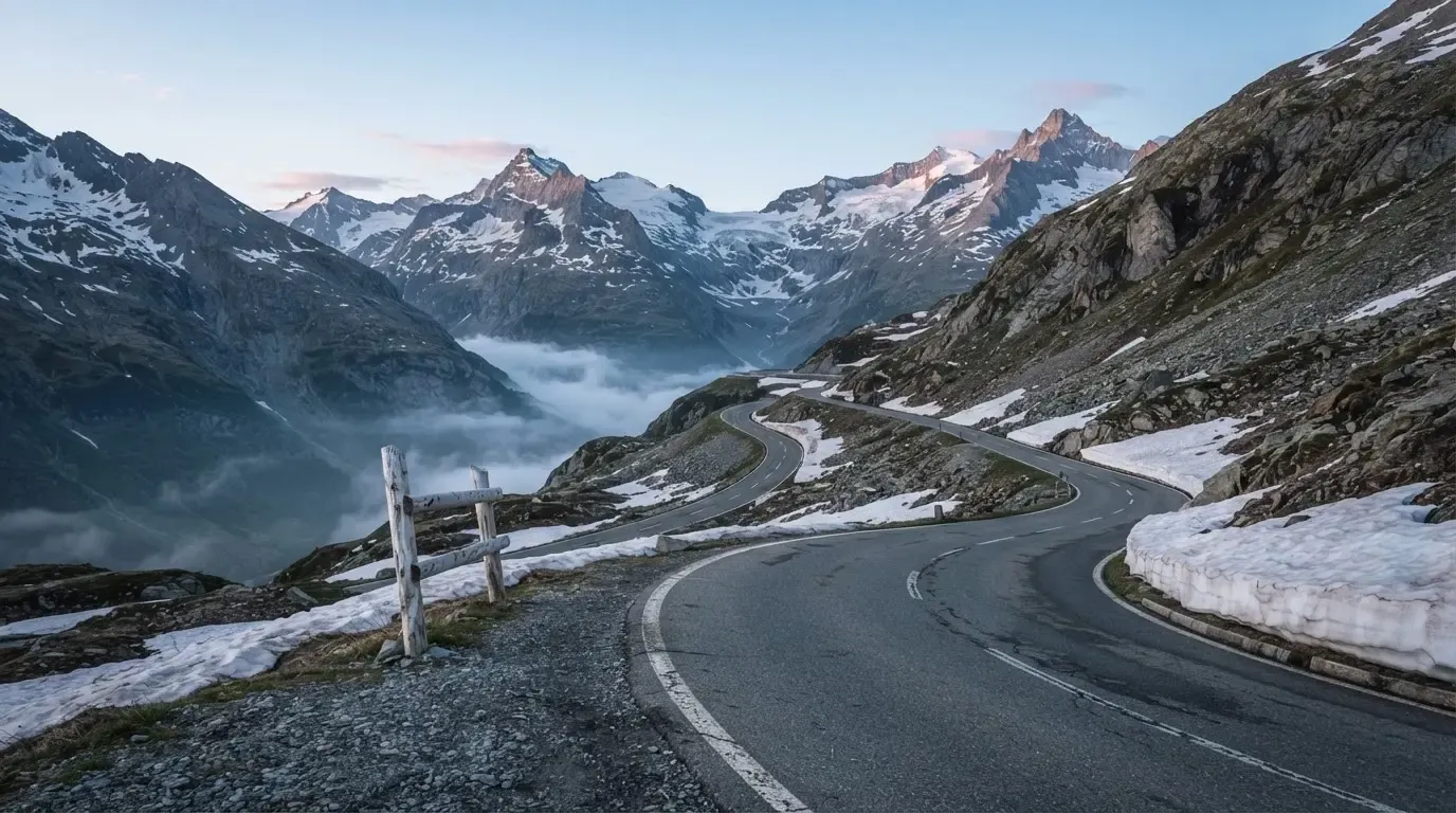 Ein Bergpass in den Schweizer Alpen mit leerer Strasse und Schneeresten am Rand im ersten Frühlicht.