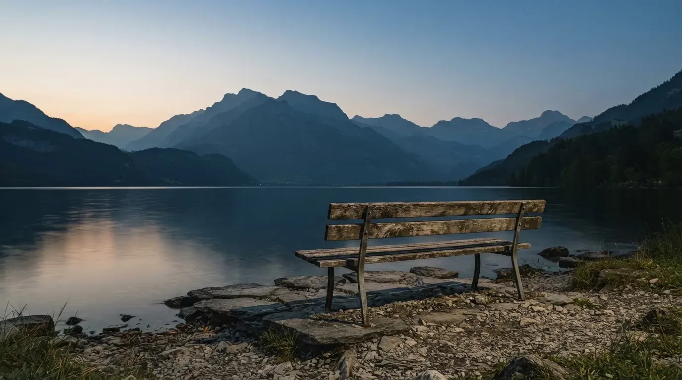 Eine leere Holzbank am Seeufer in der Dämmerung, im Hintergrund dunkle Berge