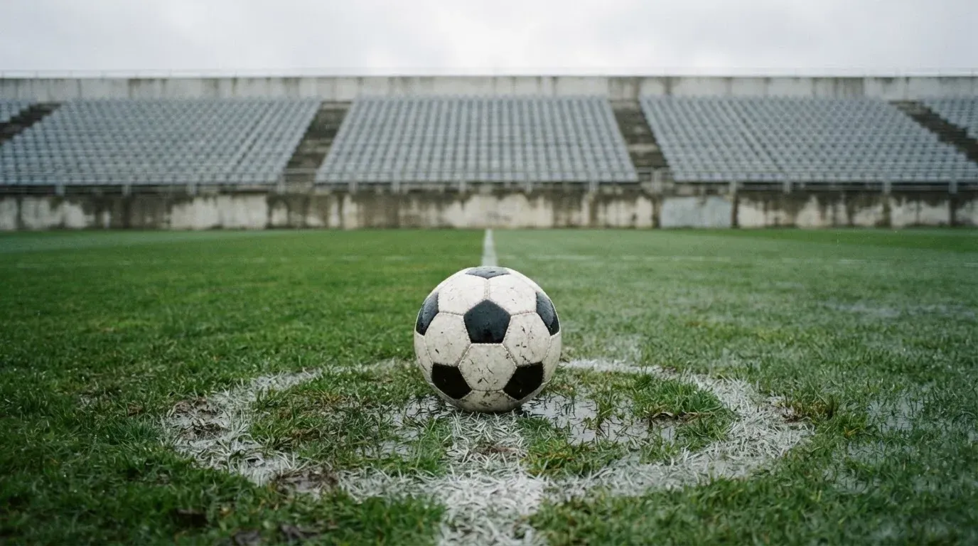 Ein Fussball liegt auf dem Anstosspunkt eines nassen Rasens, dahinter eine leere Tribüne unter grauem Himmel.