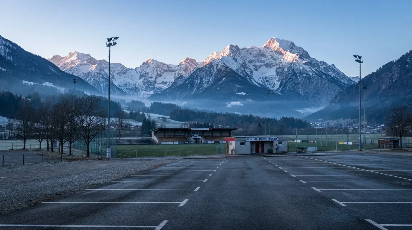 Schneebedeckte Berge ragen hinter einem kleinen Stadion in einem Alpental auf, im Vordergrund ein leerer Parkplatz am Morgen.