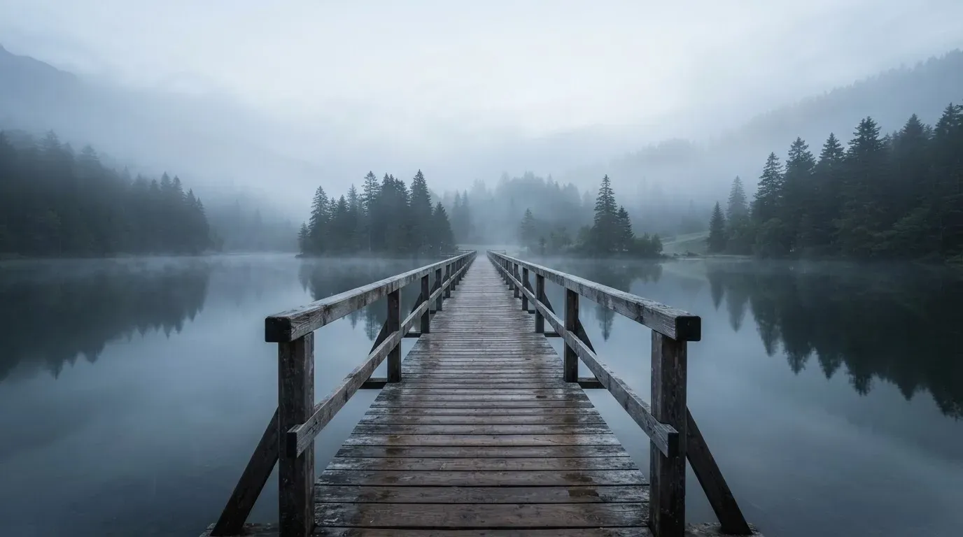 Ein Holzsteg führt über einen ruhigen Bergsee im Nebel, am anderen Ufer stehen dunkle Tannen.