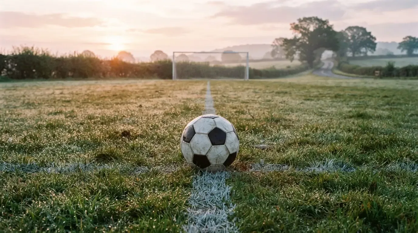 Ein Fussball liegt im Tau auf der Mittellinie eines Dorfplatzes bei Sonnenaufgang, der Rasen glitzert.