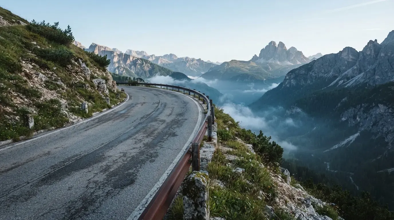 Eine Passstrasse in den Alpen mit enger Kurve und Leitplanke, darüber ein klarer Morgenhimmel und einzelne Nebelschwaden im Tal.