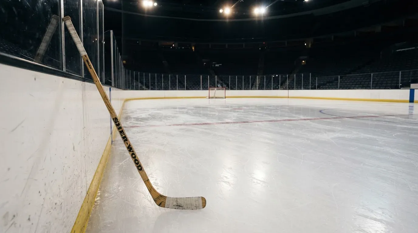 Ein Eishockeyschläger lehnt an der Bande, in der Ferne liegt die hell beleuchtete Eisfläche einer Halle unter Flutlicht.