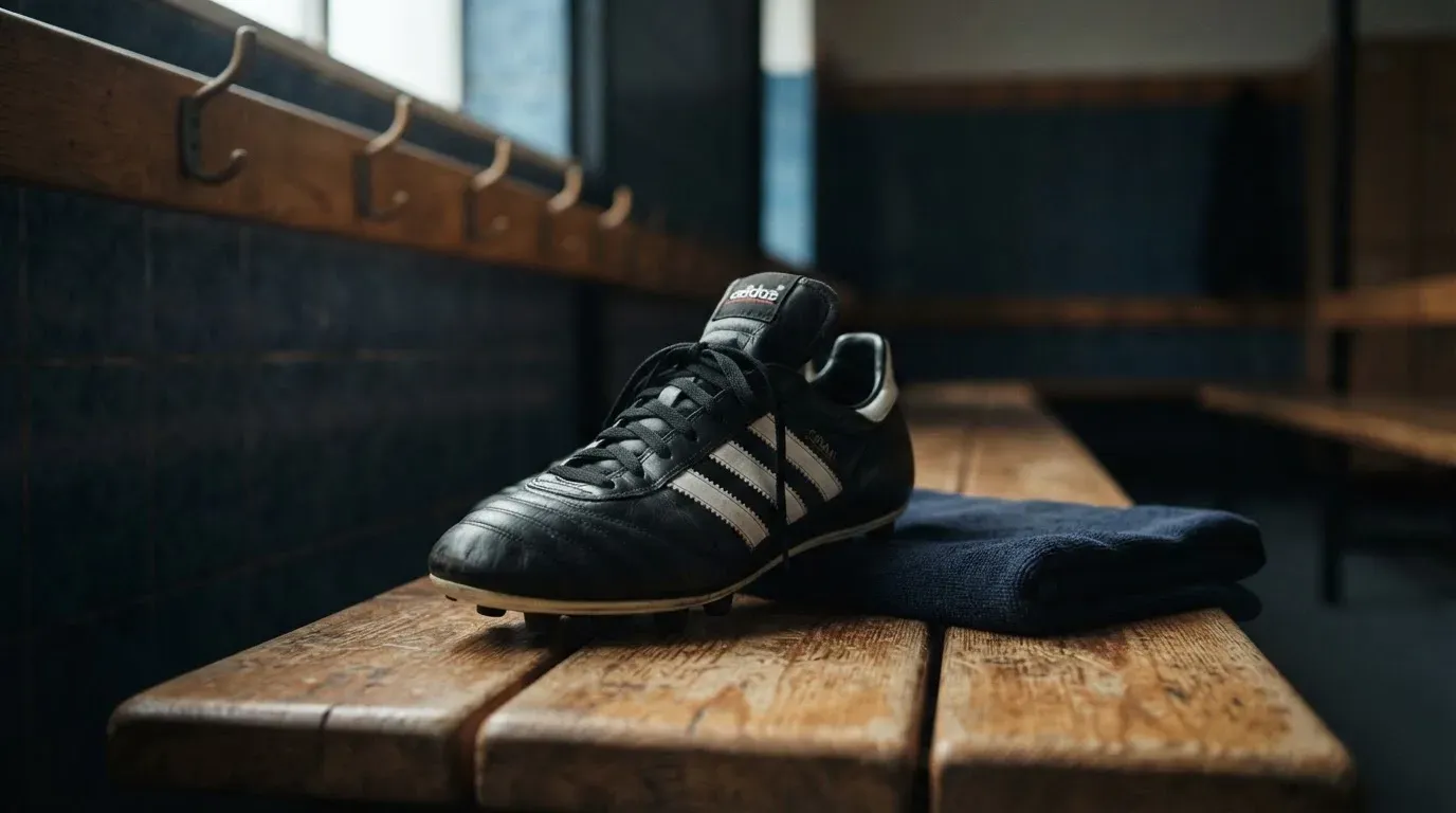 Ein Fussballschuh steht auf einer Holzbank in einer leeren Kabine, daneben liegt ein zusammengefaltetes Handtuch im Tageslicht.