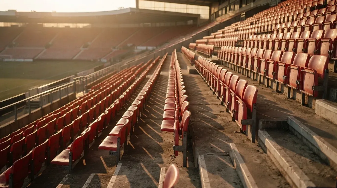 Eine Reihe leerer Sitzschalen steigt stufenweise auf einer Stadiontribüne an, die tiefe Sonne wirft lange Schatten über die Reihen.