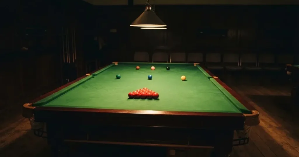 Ein Snookertisch mit grünem Tuch und einem Dreieck aus roten Kugeln unter einer tief hängenden Lampe in einer leeren Halle.