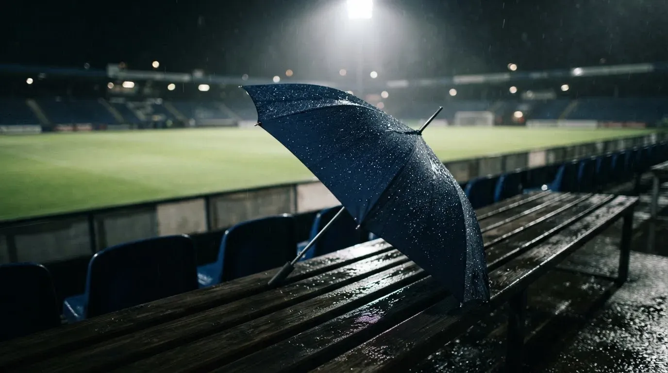 Ein aufgespannter Regenschirm liegt auf einer leeren Tribünenbank, Regentropfen glitzern im Licht der Flutlichter.