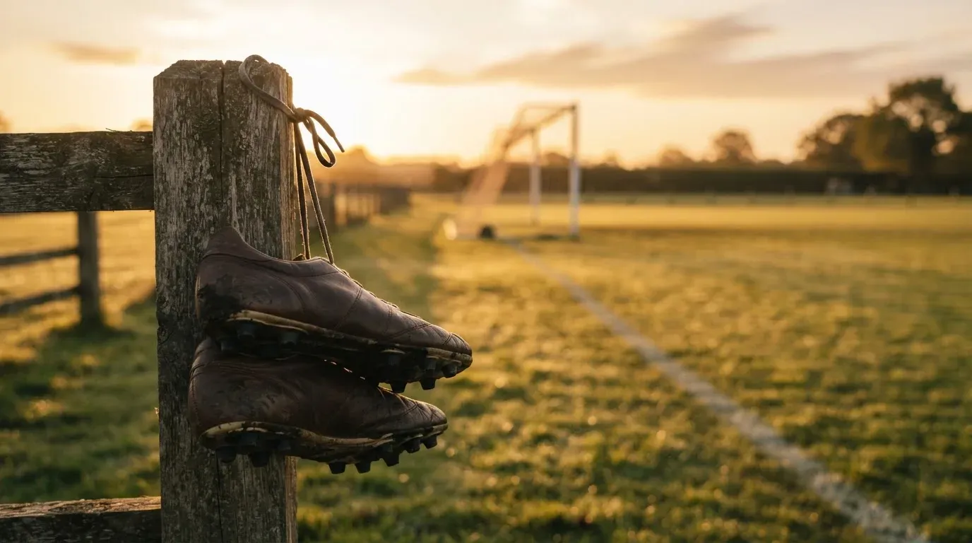 Ein Fussballschuh hängt an den Schnürsenkeln an einem Zaunpfosten neben einem Trainingsplatz im Abendlicht.