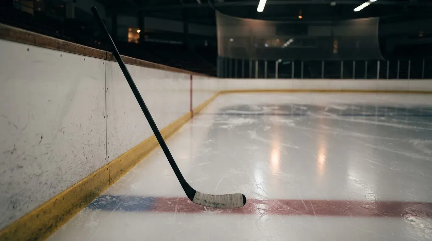 Ein Eishockeyschläger lehnt an der Bande einer leeren Halle, auf dem Eis spiegeln sich die Deckenlampen.