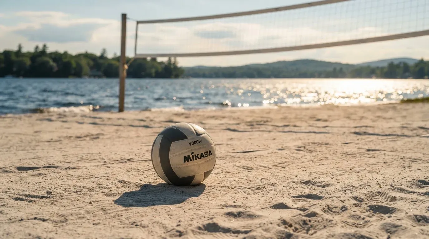 Ein Volleyball liegt auf dem Sand eines Strandfeldes am Seeufer, im Hintergrund glitzert das Wasser.