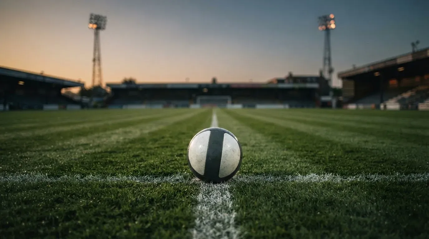 Ein Fussball liegt auf dem Mittelpunkt eines leeren Rasenplatzes, im Hintergrund ragen Flutlichtmasten in die Abenddämmerung