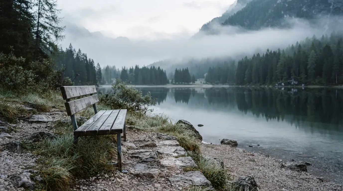Eine Holzbank am Ufer eines ruhigen Bergsees, am gegenüberliegenden Ufer steigen dunkle Hänge in den Frühnebel