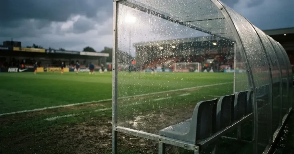 Eine leere Ersatzbank unter einem Plexiglasdach am Spielfeldrand, Regentropfen auf der Scheibe, dahinter ein Rasenplatz