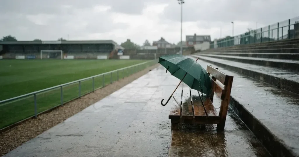 Ein offener Regenschirm lehnt an einer leeren Tribünenbank, Regen fällt auf die Stufen eines Stadions
