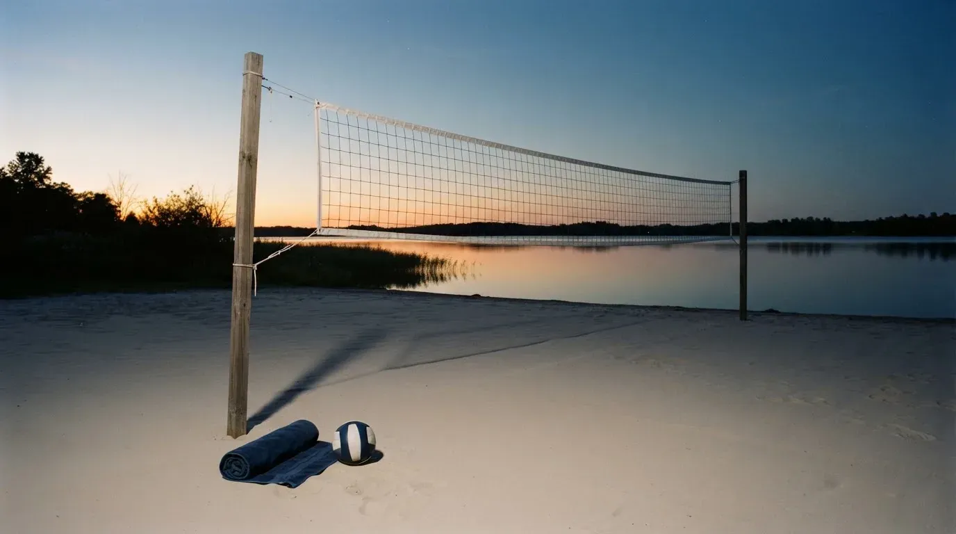 Ein Volleyballnetz steht an einem Seeufer in der Abenddämmerung, der Sand ist leer und das Wasser glatt.