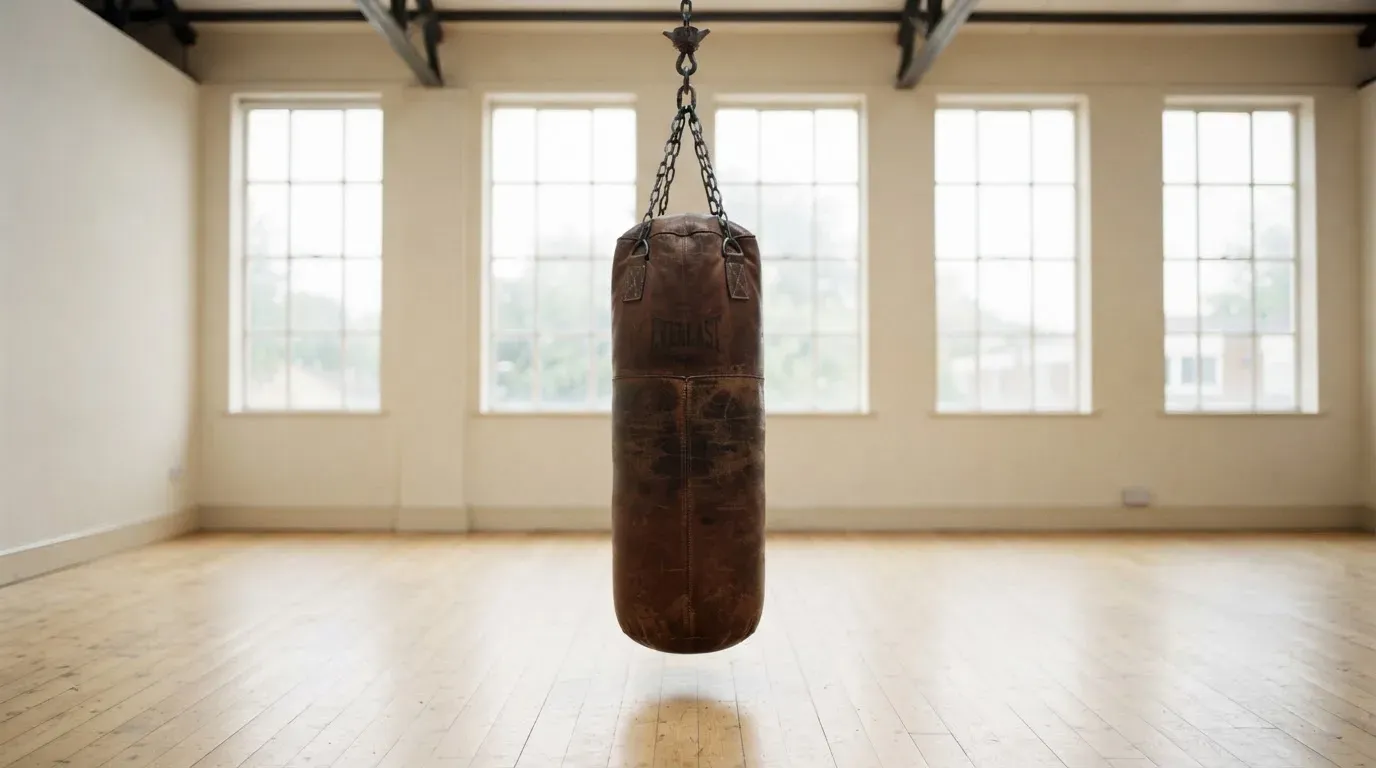 Ein Boxsack hängt an einer Kette in einem leeren Trainingsraum mit hellem Holzboden und grossen Fenstern.