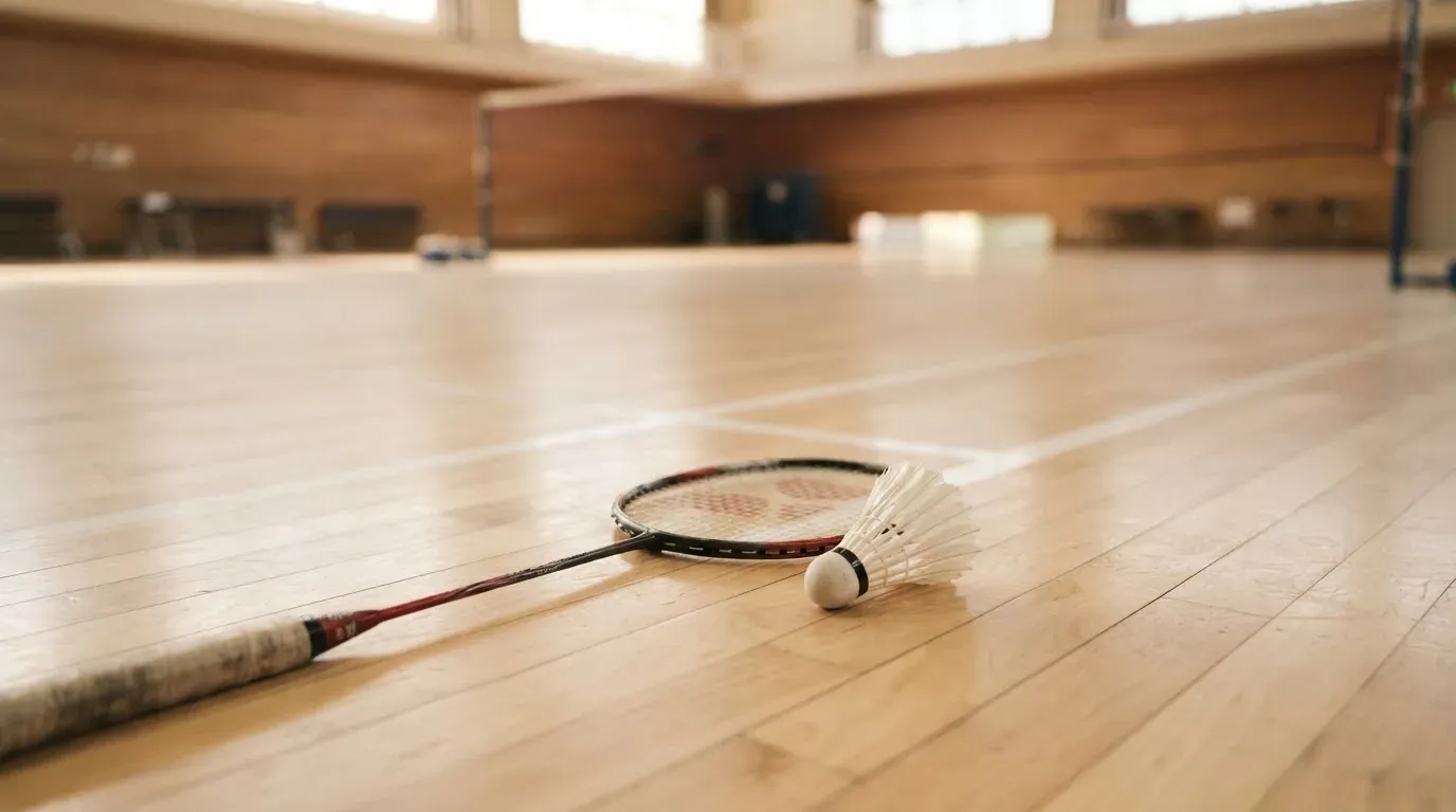 Ein Badmintonschläger liegt neben einem Federball auf dem hellen Hallenboden eines leeren Spielfelds.