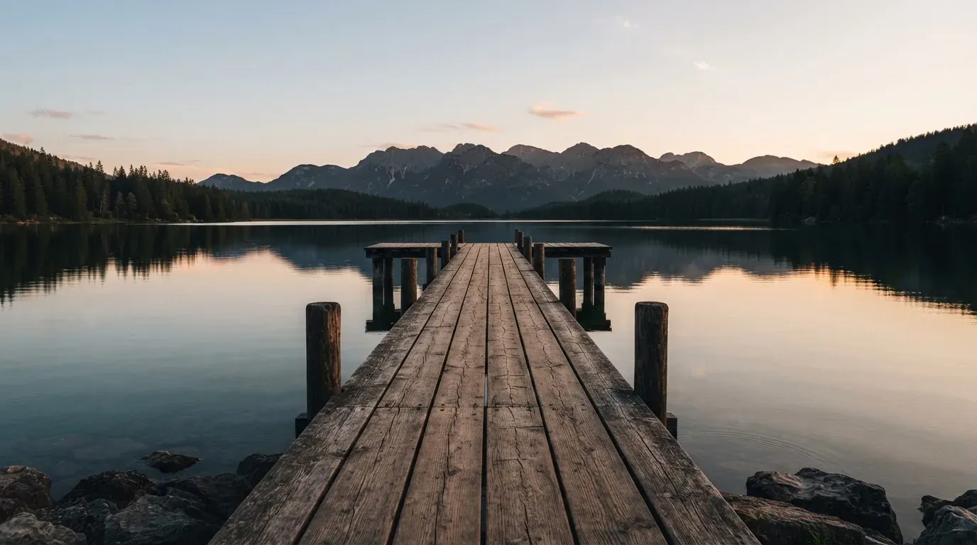 Ein leerer Holzsteg ragt am Abend in einen ruhigen Bergsee, das Wasser spiegelt den Himmel.