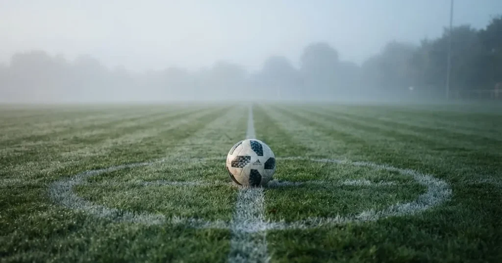 Ein Fussball liegt auf dem Anstosspunkt eines leeren Rasenplatzes im dichten Morgennebel.