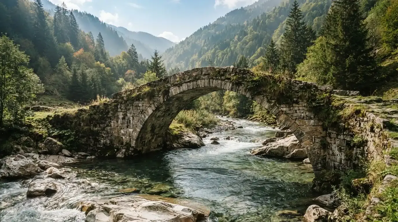 Eine alte Steinbrücke überspannt einen Gebirgsfluss, im Hintergrund steigen bewaldete Hänge an.