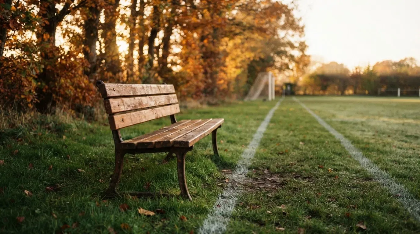 Eine hölzerne Bank steht an der Seitenlinie eines leeren Sportplatzes, dahinter Bäume im Herbstlicht.